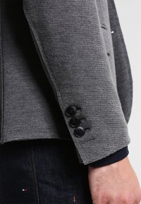 Harmaa bleiseri, jossa on teksturoidusta kankaasta tehty, kolmen napin kalvosimet, terävät kaulukset ja tyylikäs istuvuus. Koristeena mustat napit.