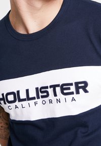 Mörkblå crewneck t-shirt med en vit horisontell rand och broderad text "HOLLISTER CALIFORNIA" i marinblått. Tillverkad av bomull med en slät textur.