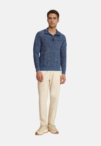 Dunkelblauer Strickpullover mit einem gestreiften Kragen, halbem Reißverschluss und strukturiertem Muster, kombiniert mit beigen Hosen und hellen Sneakers.