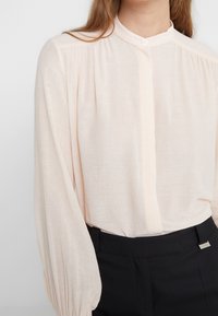 Světle růžová blůza s dlouhým rukávem a mandarínkovým límcem, zapínání na knoflíky, měkká textura a nabírané rukávy, v kombinaci s černými krejčovskými kalhotami.