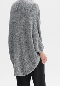 Asymetryczny szary sweter z dzianiny o fakturowanym wykończeniu, luźnym kroju i długich rękawach, zestawiony z czarnymi spodniami.