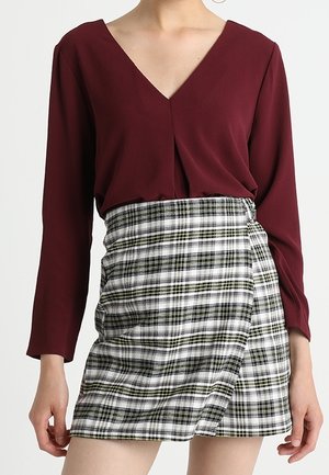 Haut bordeaux à manches longues avec un col en V, associé à une jupe portefeuille à carreaux noir et blanc avec des accents verts et un détail noué sur le côté.