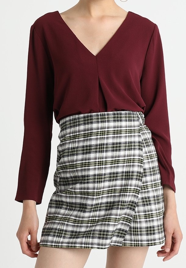 Top bordeaux a maniche lunghe con scollo a V, abbinato a una gonna avvolgente in plaid bianco e nero con dettagli verdi e nodo laterale.