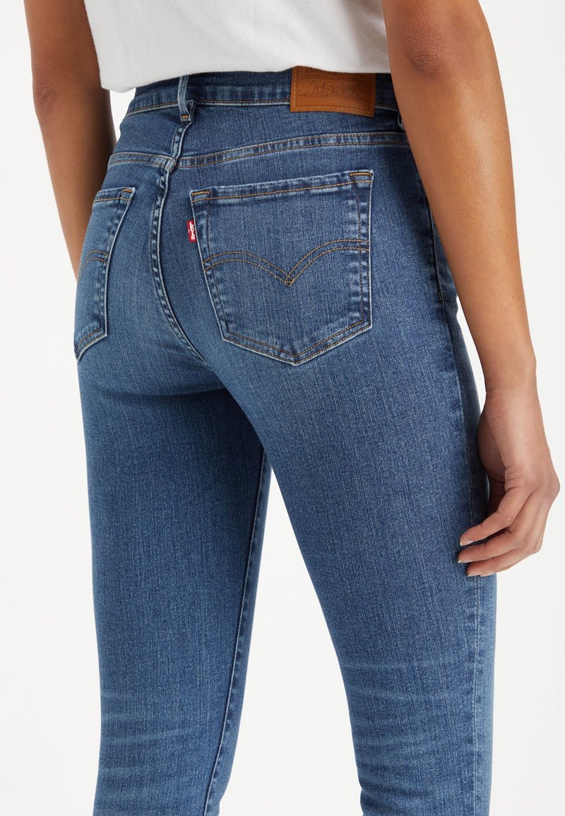 Jeans en denim bleu avec une taille haute, présentant des poches arrière distinctives et une patch en cuir marron à la taille.