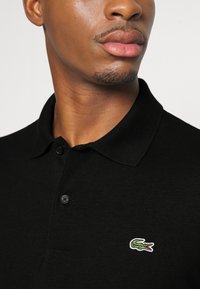 Černé polo triko s texturovaným povrchem, s dvouknoflíkovým zapínáním a zeleným vyšívaným krokodýlem na levé straně hrudníku.