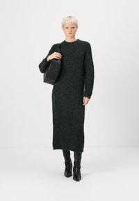 Καστόν φόρεμα σε σκούρο πράσινο με μακριά μανίκια και χαλαρή εφαρμογή, κατασκευασμένο από μαλακό και αφράτο υλικό, συνδυασμένο με μαύρες μπότες μέχρι το γόνατο και μαύρη τσάντα.