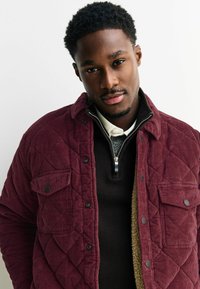 Bordeaux gekoord quilted jas met drukknoopsluitingen, voorzien van een afneembare kraag en een bruine trui eronder, weergegeven tegen een effen achtergrond.