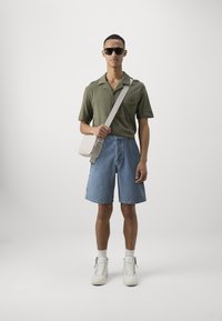 Chemise verte à manches courtes, shorts en denim bleu clair, baskets montantes blanches et sac bandoulière gris avec sangle réglable. Lunettes de soleil noires.