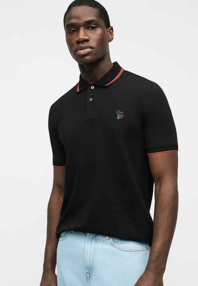 Νεαρός άνδρας με μαύρο πόλο πουκάμισο με πορτοκαλί λεπτομέρειες και μικρό πολύχρωμο λογότυπο, συνδυασμένο με ανοιχτό μπλε τζιν, στεκόμενος μπροστά σε λευκό φόντο.