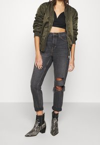 Olive green bomber jacket over a black cropped top, paired with high-waisted gray jeans featuring knee rips and snake-print ankle boots.