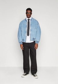 Light blue denim jacket with ribbed cuffs, worn over a white shirt and black tie, paired with black wide-leg trousers and black sneakers.