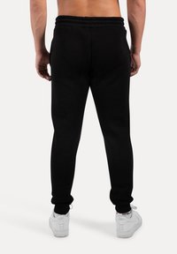 Sorte sweatpants lavet af blød stof, med elastisk talje og mansjetter, tapering design og minimalistisk stil. Hvide sneakers synlige.
