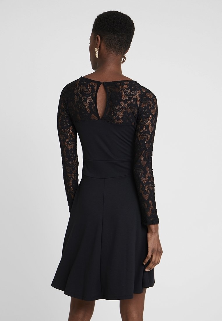 Femme portant une robe noire avec des manches longues en dentelle et un détail en trou de serrure dans le haut du dos, debout devant un fond uni.