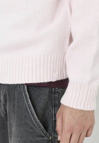 Pull tricoté rose clair avec poignets et ourlet côtelés. Présente une texture douce et une petite étiquette en tissu sur le côté. Porté avec un pantalon gris foncé.