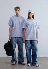 Ανοιχτό μωβ βαμβακερά μπλουζάκια, άνετη εφαρμογή. Συνδυάζονται με ξεβαμμένο μπλε τζιν τζιν, ένα άτομο κρατάει μια μαύρη τσάντα duffel, ενώ το άλλο φοράει ροζ καπέλο.