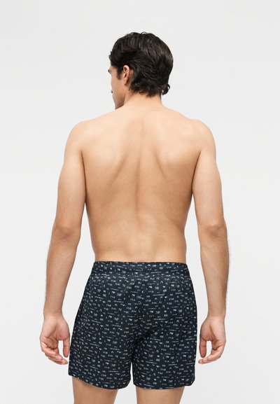 Άνδρας με γυμνή πλάτη, φορώντας μαύρο μαγιό με λευκά γράμματα, στέκεται μπροστά σε απλό ανοιχτό γκρι φόντο.