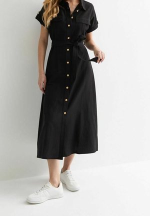 Femme portant une robe midi noire à boutons sur le devant, manches courtes et baskets blanches, tenant une ceinture à nouer à la taille.