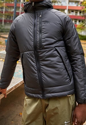 Homme portant une doudoune noire Nike et un short beige Nike, debout à l'extérieur près d'une table en métal avec une vue sur la ville en arrière-plan.