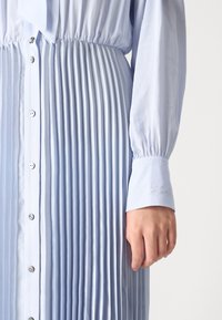 Vestido pregueado azul claro com frente de botões e cintura franzida. Apresenta mangas longas e detalhe de punho bordado. Textura de tecido suave.