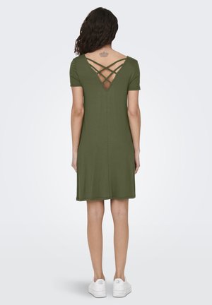 Mujer con vestido de manga corta color verde oliva con detalle en la espalda en forma de cruz, de pie de espaldas, combinando con zapatillas blancas.