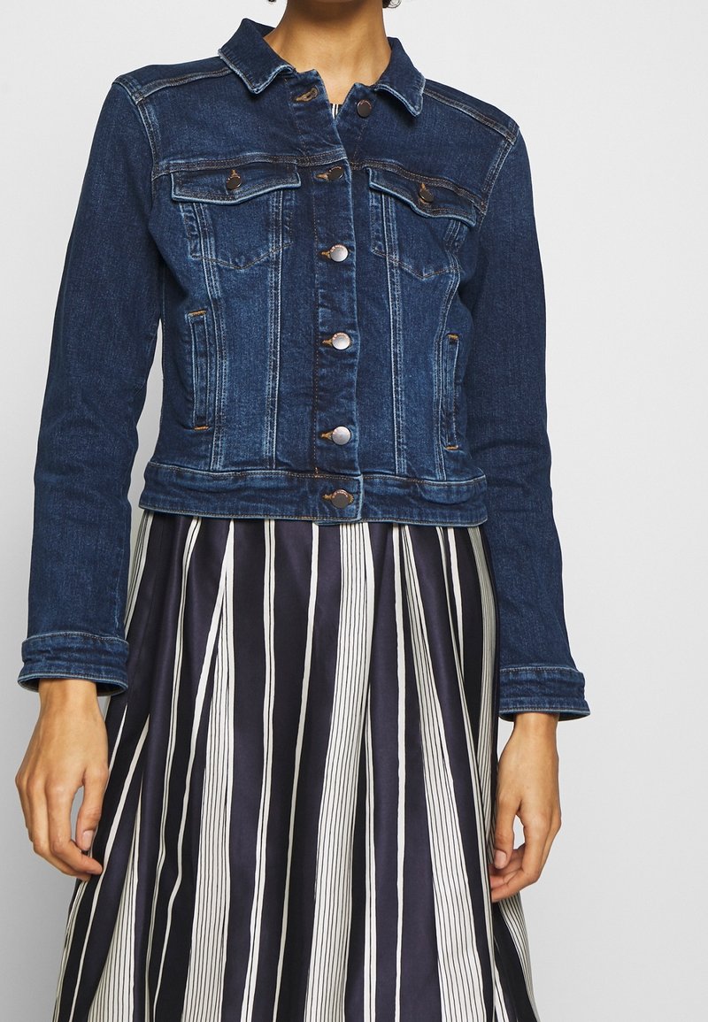 Dark denim jacket with button front, two chest pockets, and a fitted design. Worn over a navy and white striped skirt.