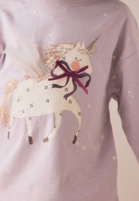Pull lavande pour enfant avec une application décorative licorne comprenant un nœud violet, des ailes en tissu et des motifs d’étoiles en arrière-plan.