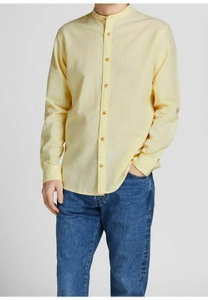 Man met een lichtgeel overhemd met mandarijnkraag en knopen, lange mouwen en een blauwe spijkerbroek, met één hand in de zak, neutrale achtergrond.