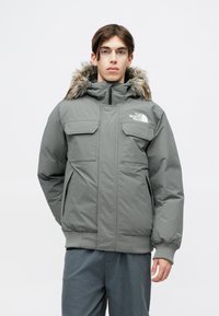 Graue isolierte Jacke mit einem mit Fell gefütterten Kapuzen, ausgestattet mit zwei Brusttaschen und gerippten Bündchen. Logo auf der linken Brust. Glatte, matte Oberfläche.