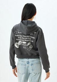 Sudadera corta gris con capucha ajustable. Presenta texto blanco y diseños gráficos que incluyen estrellas, fresas y elementos textuales en la parte posterior.