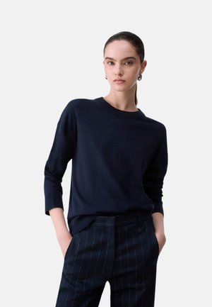 Donna con capelli scuri raccolti in una coda bassa, indossa un maglione blu navy a maniche lunghe e pantaloni a righe blu navy, mani in tasca, sfondo neutro.