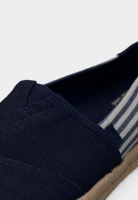 Μπλε ναυτικό πάνινο παπούτσι χωρίς κορδόνια με ραβδωτό ύφασμα στην πλάτη και φυσική πλεγμένη σόλα, κοντινή λήψη που δείχνει την υφή και τις λεπτομέρειες των ραφών.