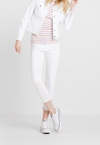 Giacca di denim bianca sopra una maglietta a righe rosse e bianche, abbinata a pantaloni bianchi alla caviglia e sneakers bianche. Design semplice, vestibilità casual.