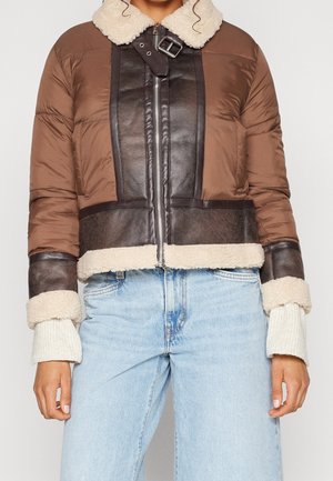 Brun og mørk læderjakke med lammeskindskrave og manchetter, båret med lyseblå jeans og synlige hvide strikkede ærmer.