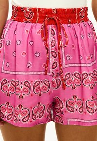 Pantalones cortos rosas con un cinturon fruncido rojo, que presentan diseños de paisley y corazones. Los lazos en la parte delantera añaden un detalle al estilo casual.
