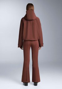 Hnedá mikina s kapucňou a vysokým golierom, kombinovaná s priliehavými rozšírenými nohavicami s všitými zipsami na členkoch. Mäkko textúrovaná tkanina.