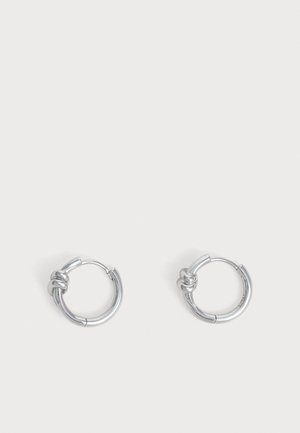 Paire de petites boucles d’oreilles créoles en argent, chacune ornée d’un détail de nœud unique sur fond gris clair.