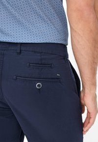 Pantaloni blu navy in cotone con una superficie liscia, dotati di due tasche posteriori, una con chiusura a bottone e dettagli di cucitura discreti.