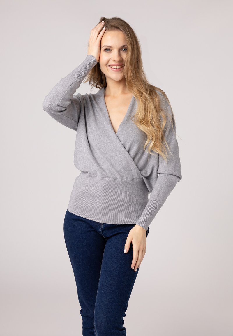 Grijze wikkeltrui met een diepe V-hals en lange mouwen, voorzien van een geribbelde zoom en een zachte textuur, gecombineerd met donkerblauwe jeans.