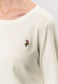 Crèmekleurige gebreide trui met een brede halslijn. Voorzien van een klein geborduurd logo op de borst in meerkleurige draad. Zachte textuur.