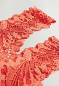 Koraalkleurige kantstof met ingewikkelde bloemenpatronen, gegolfde randen en een gestructureerd oppervlak, geschikt voor kleding of versieringen.
