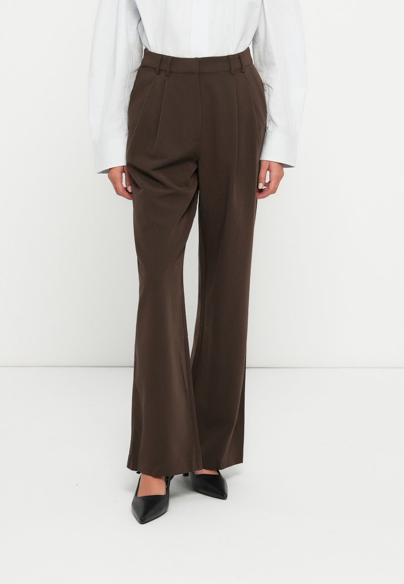 Pantaloni marroni a vita alta con pieghe, gamba ampia e una texture liscia, abbinati a décolleté nere a punta.