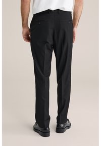 Svarta skräddarsydda byxor med slät front, två bakfickor och en smal passform. Bärs med svarta skor mot en neutral bakgrund.