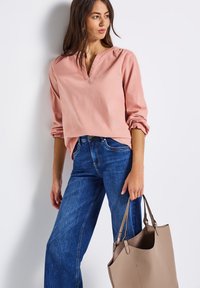 Helles rosa Langarmbluse mit V-Ausschnitt, kombiniert mit blauen Jeans. Glatte Textur, elastische Bündchen und eine minimalistische sandfarbene Tragetasche.