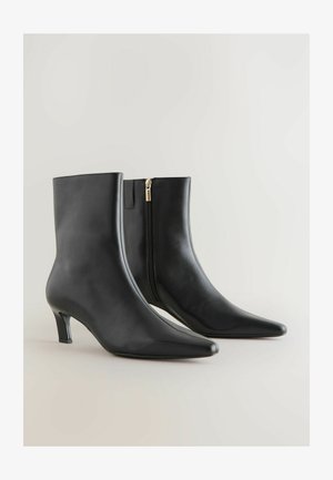 Crne kožne gležnjače s uskim vrhom i malim potpeticama. Imaju bočni zatvarač s zlatnim povlačnim dijelom. Glatka tekstura, elegantan dizajn.