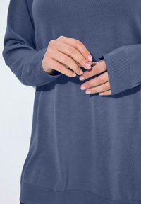 Blauer gestrickter Pullover mit gerippten Bündchen, der eine lockere Passform und eine weiche Textur aufweist, dargestellt mit Händen, die den Ärmel anpassen.
