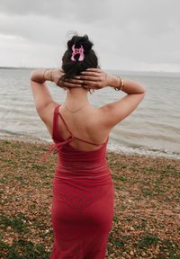 Vékony pántos, piros pöttyös ruha, testhezálló szabással és fodros textúrával. Egy rózsaszín hajcsat díszíti a konty frizurát. A háttérben vízpart látható.