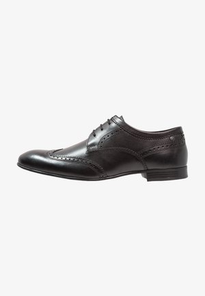 Chaussure brogue en cuir noir avec un bout arrondi, des perforations détaillées et six œillets pour les lacets. Texture lisse, talon empilé bas.