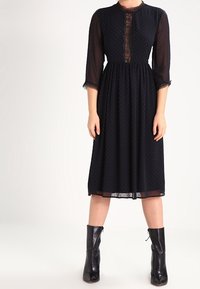 Schwarzes Kleid mit durchsichtigen langen Ärmeln, gepunktetem Muster, Rundhalsausschnitt und Spitzenakzenten vorne. Midi-Länge und tailliert. Schwarze Stiefel.