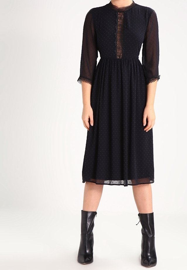 Schwarzes Kleid mit durchsichtigen langen Ärmeln, gepunktetem Muster, Rundhalsausschnitt und Spitzenakzenten vorne. Midi-Länge und tailliert. Schwarze Stiefel.