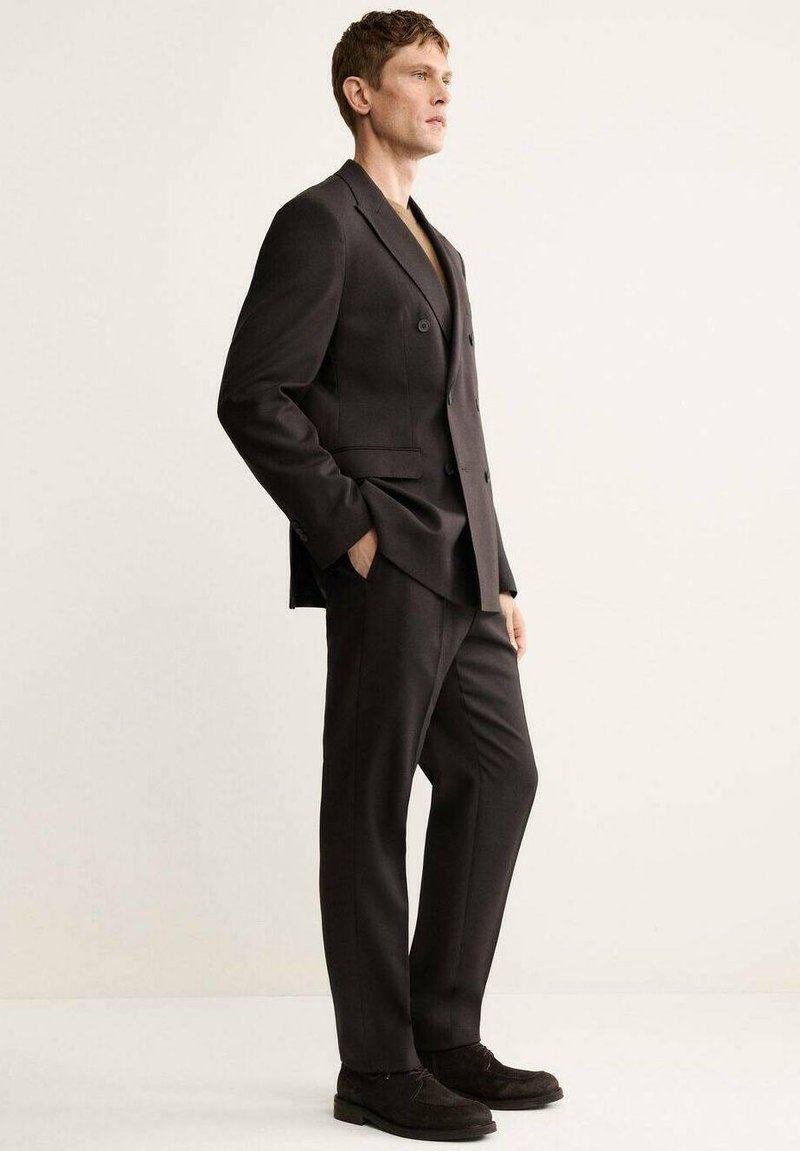 Dark brown, double-breasted suit with a tailored fit, accompanied by a dark undershirt and black suede shoes, standing against a neutral background.